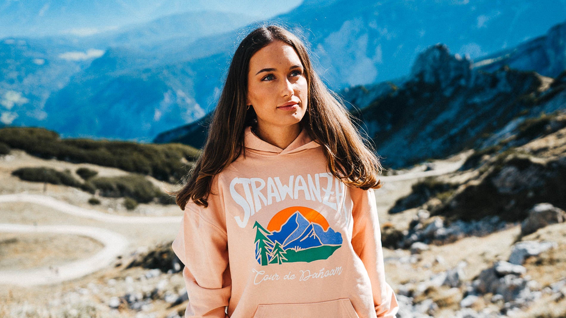 Junge Frau präsentiert einen Kapuzenpullover der Modemarke Strawanza in einer Berglandschaft.