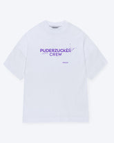 Ein weißes T-Shirt mit einem lilanen Schriftzug auf der Vorderseite welcher "PUDERZUCKER CREW" lautet, wobei des Ende des ersten Wortes sich in kleine Splitter auflöst. 