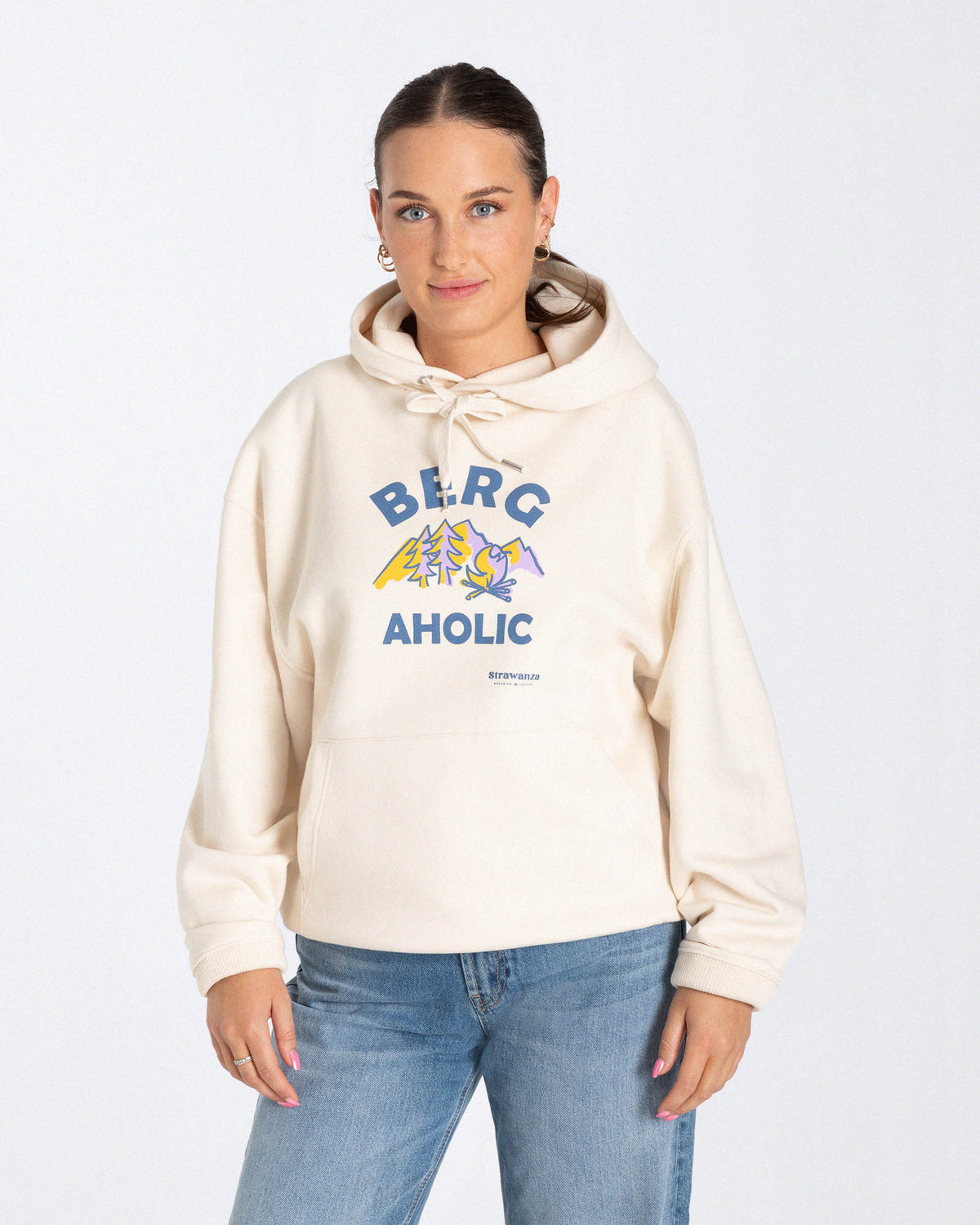 Ein weibliches Model trägt einen Hoodie in der Farbe Natur mit einem Motiv vorne auf der Höhe der Brust, welches aus lila-orangenen Bergen, Bäumen und einem Lagerfeuer besteht und einem dunkelblauen "BERGAHOLIC" Schriftzug.