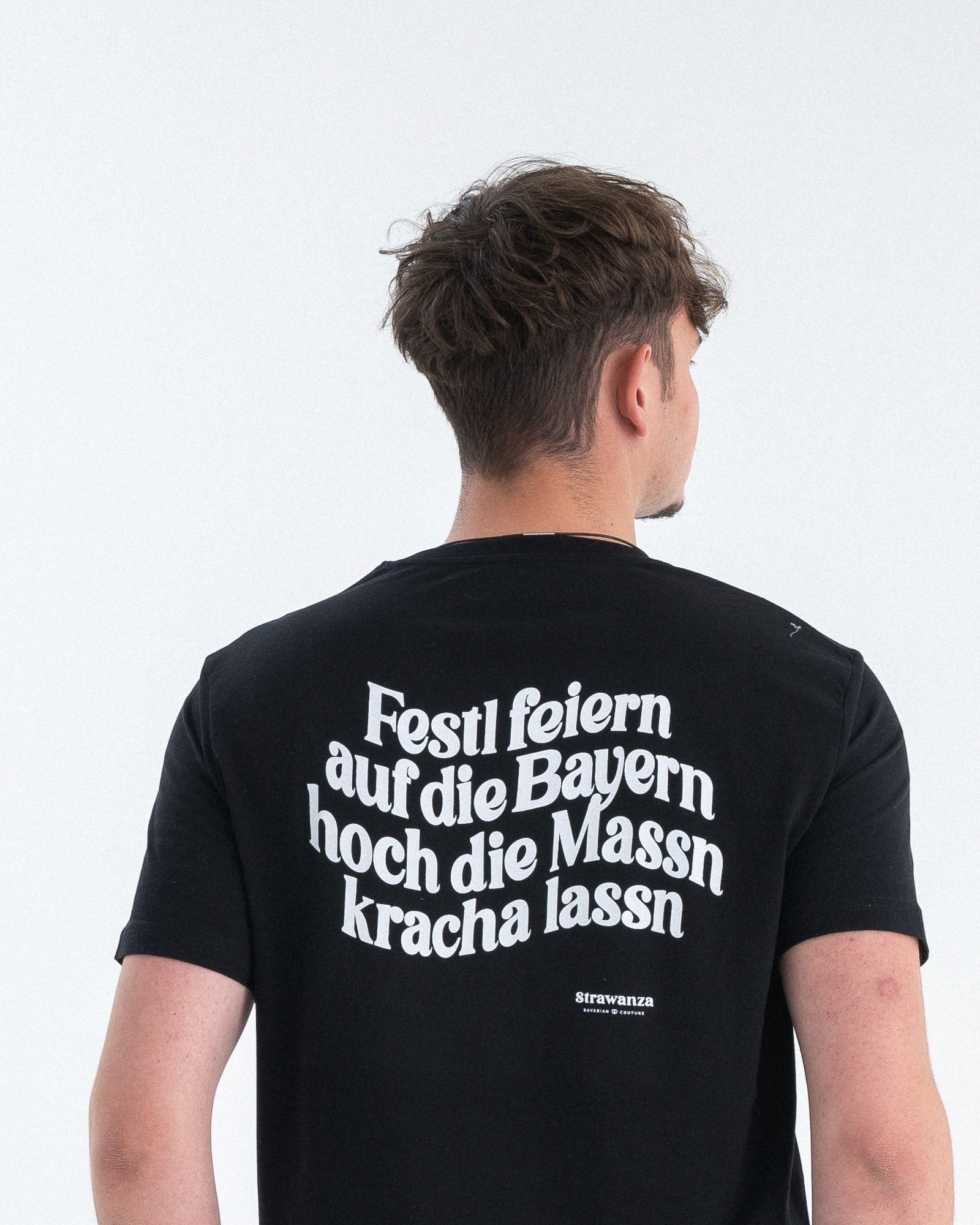 Ein männliches Model trägt ein schwarzes T-Shirt mit einem in weiß gedruckten "Festl feiern auf die Bayern hoch die Massn kracha lassn" Schriftzug. 