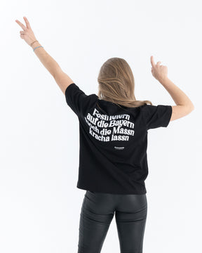 Ein weibliches Model trägt ein schwarzes T-Shirt mit einem weißen Backprint wo "Festl feiern auf die Bayern hoch die Massn kracha lassn" steht.