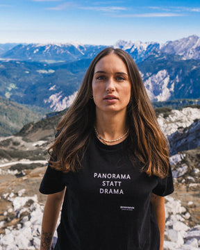 Eine Frau trägt ein schwarzes T-Shirt mit einer weißen "PANORAMA STATT DRAMA" Schrift auf der Brust in den Bergen. 
