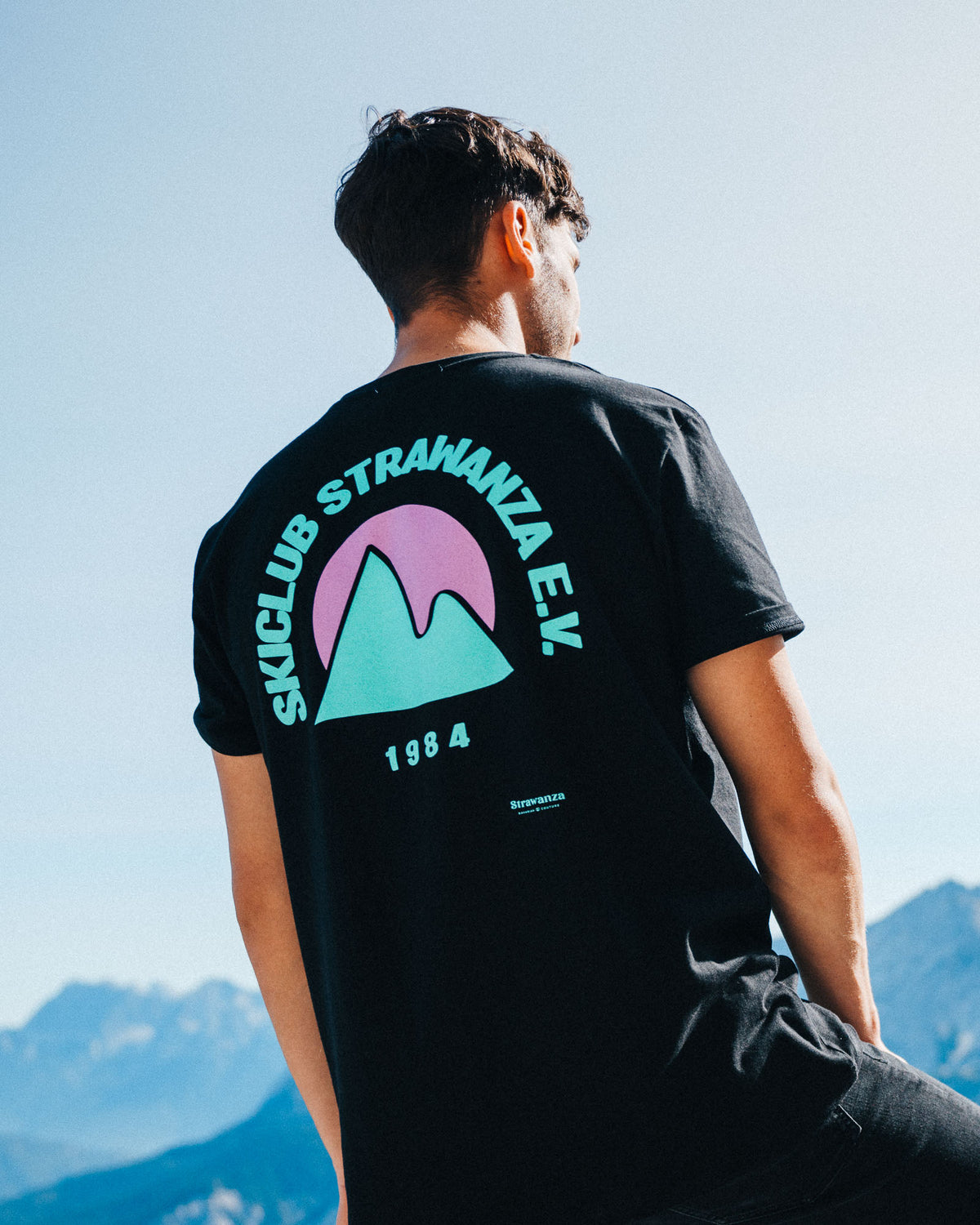 Ein männliches Model trägt ein schwarzes T-Shirt mit einem Backprint in den Bergen, was aus einem türkisen Berg, einer pinken Sonne und einem türkisen "SKICLUB STRAWANZA E.V." und "1984" Schriftzug besteht.