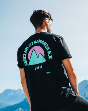 Ein männliches Model trägt ein schwarzes T-Shirt mit einem Backprint in den Bergen, was aus einem türkisen Berg, einer pinken Sonne und einem türkisen "SKICLUB STRAWANZA E.V." und "1984" Schriftzug besteht. 