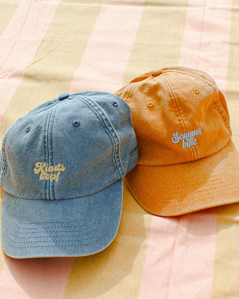 Eine Vintage orange und Vintage Light Denim Bavarian Cap liegen auf einem Strandtuch, die orange hat einen weißen "Sommer bitte" Schriftzug während die Light denim einen gelben "Kindskopf" Schriftzug auf der Vorderseite hat.