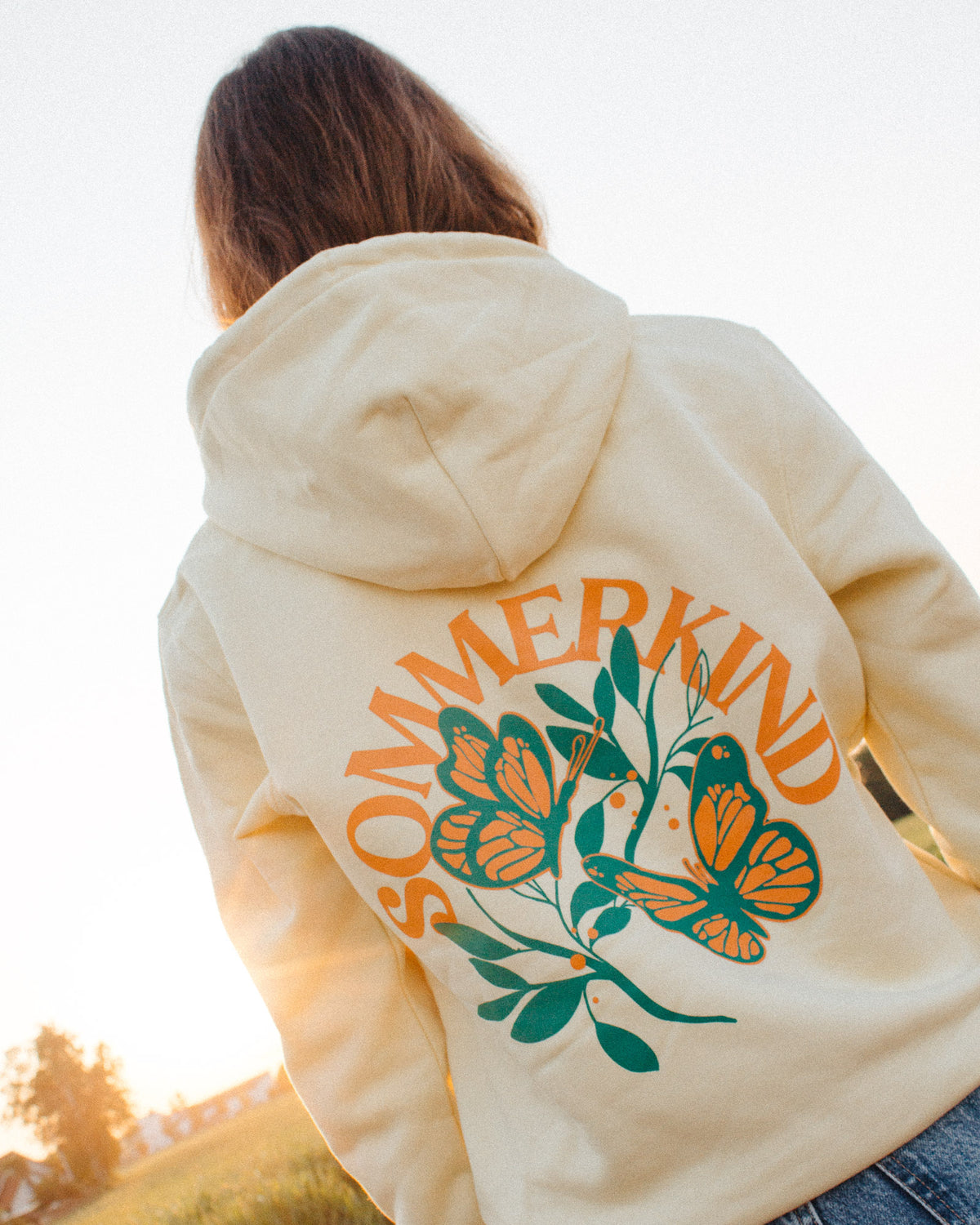Ein weibliches Model trägt einen hellgelben Hoodie mit einem Backprint, welcher aus zwei orange, grünen Schmetterlingen, einem grünen Ast und einem orangenen "SOMMERKIND" Schriftzug besteht.