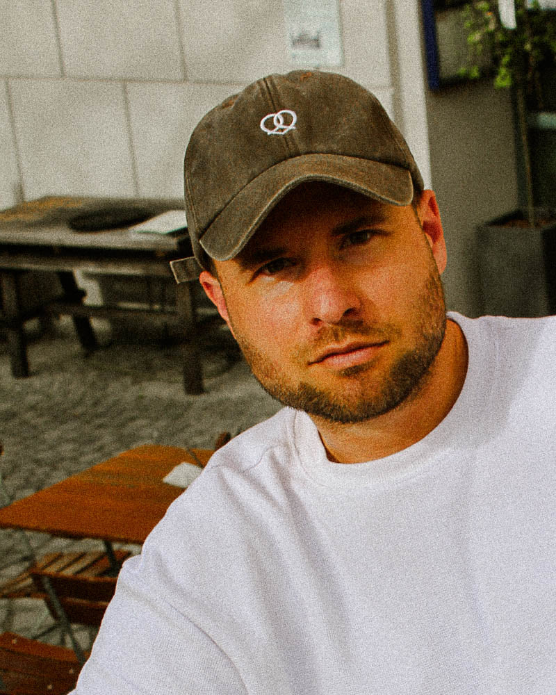 Ein männliches Model trägt eine Vintage braune Bavarian Cap mit einem Brezn-Herz Logo auf der Vorderseite der Cap in weiß.