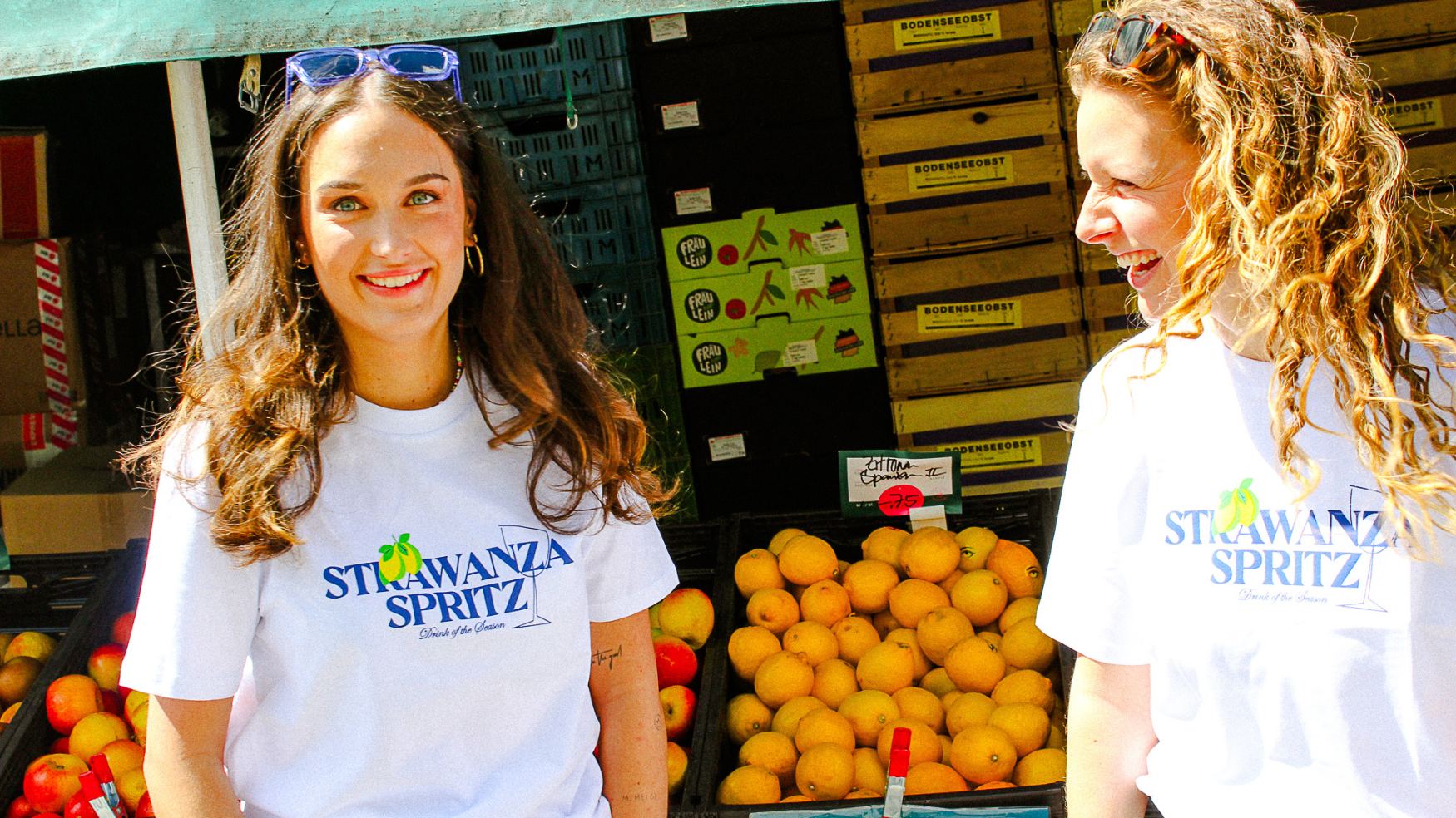 Zwei junge Frauen stehen am Viktualienmarkt in München und tragen neue Sommershirt der Modemarke Strawanza Bavarian Couture. Auf den T-Shirts sind Zitronen abgebildet. 