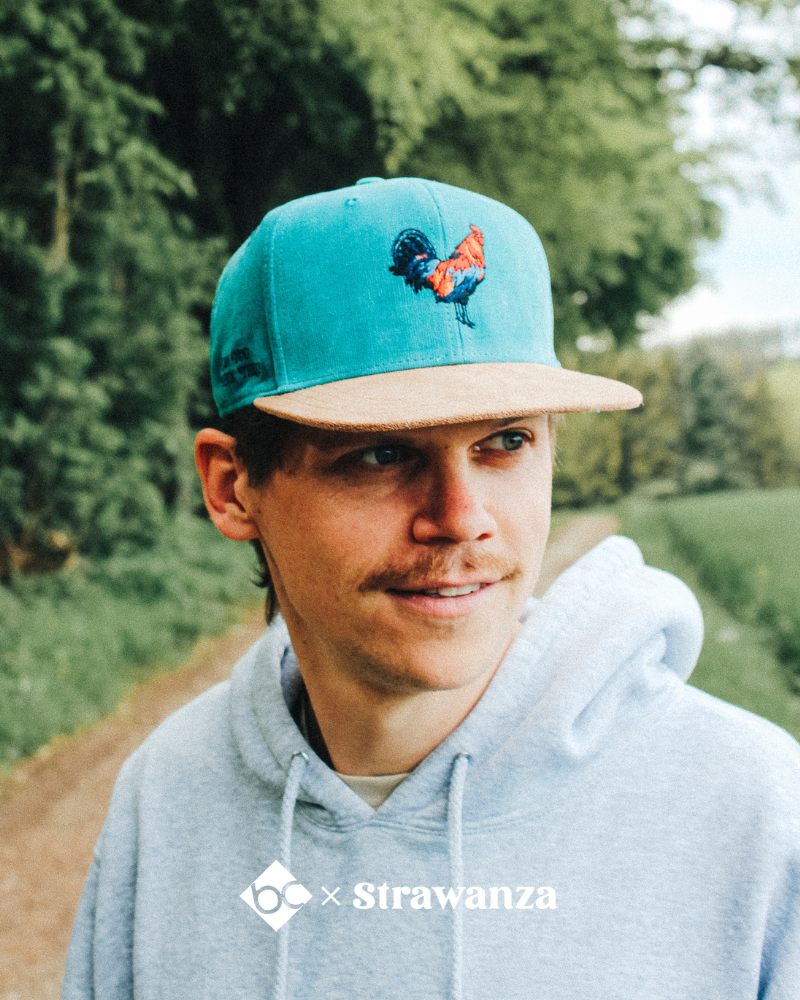 Ein männliches Model trägt eine türkise bayerische Cap mit einem "Hendl" vorne als Motiv.