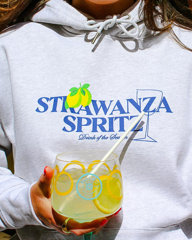 Ein weibliches Model trägt einen Hoodie in der Farbe cool hellgrau mit einem Motiv aus einer Zitrone, einem Sektglas und einem dunkelblauen "STRAWANZA SPRITZ" Schriftzug und hält währenddessen ein Getränk in der Hand.