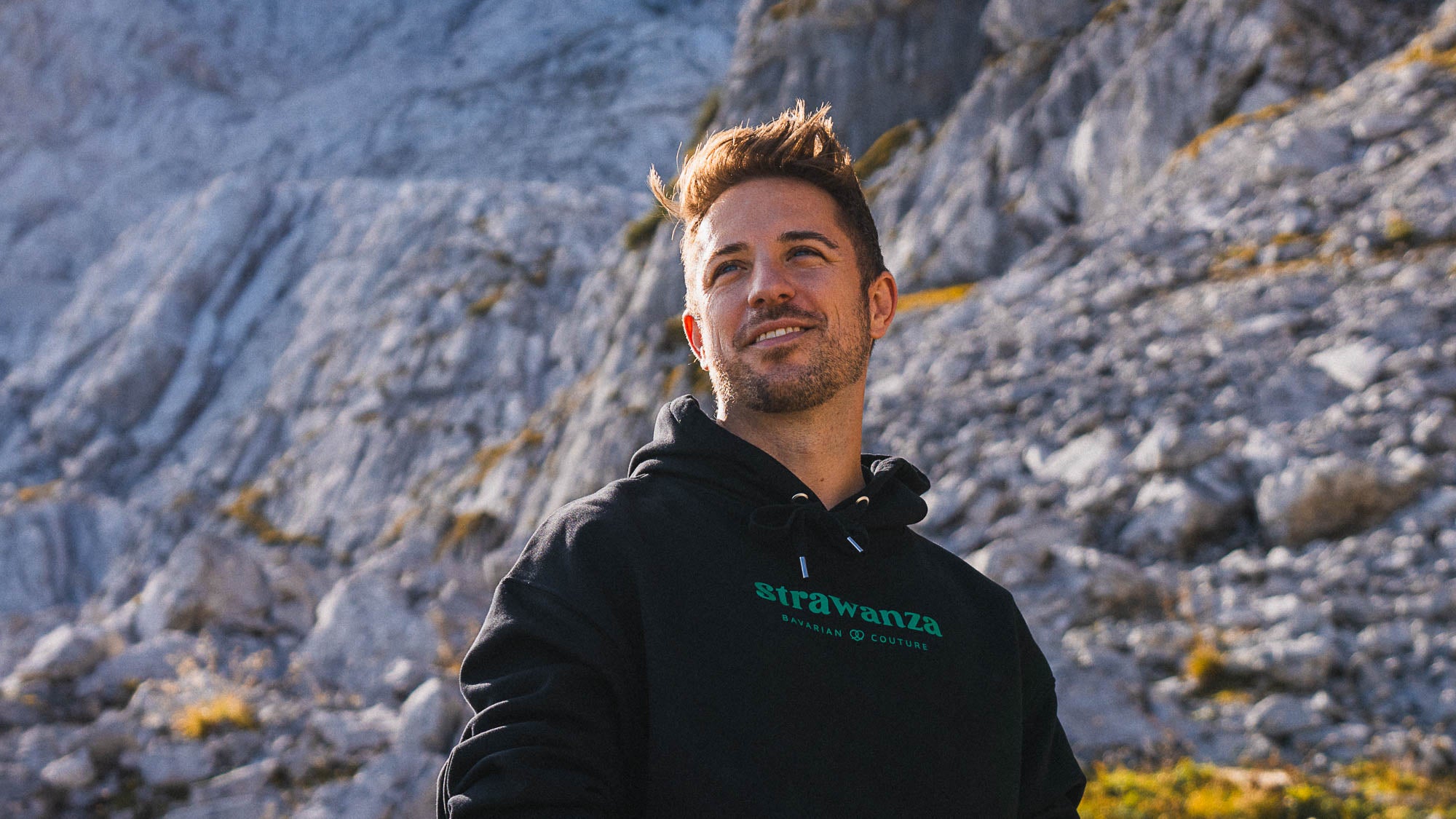 Männliches junges Model in schwarzem Sweatshirt mit grüner Bedruckung sitzt vor Bergkulisse in der bayerischen Natur. 