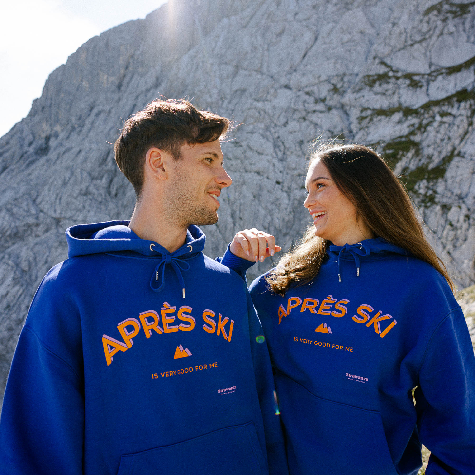 Der Après Ski Hoodie in blau zu sehen an einem Mann und einer Frau in einer Bergkulisse. 