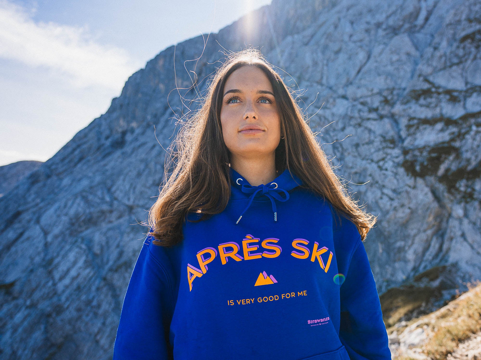 Weibliches Model trägt blauen Après Ski Hoodie in Übergröße. Zu sehen in schönem Bergpanorama.