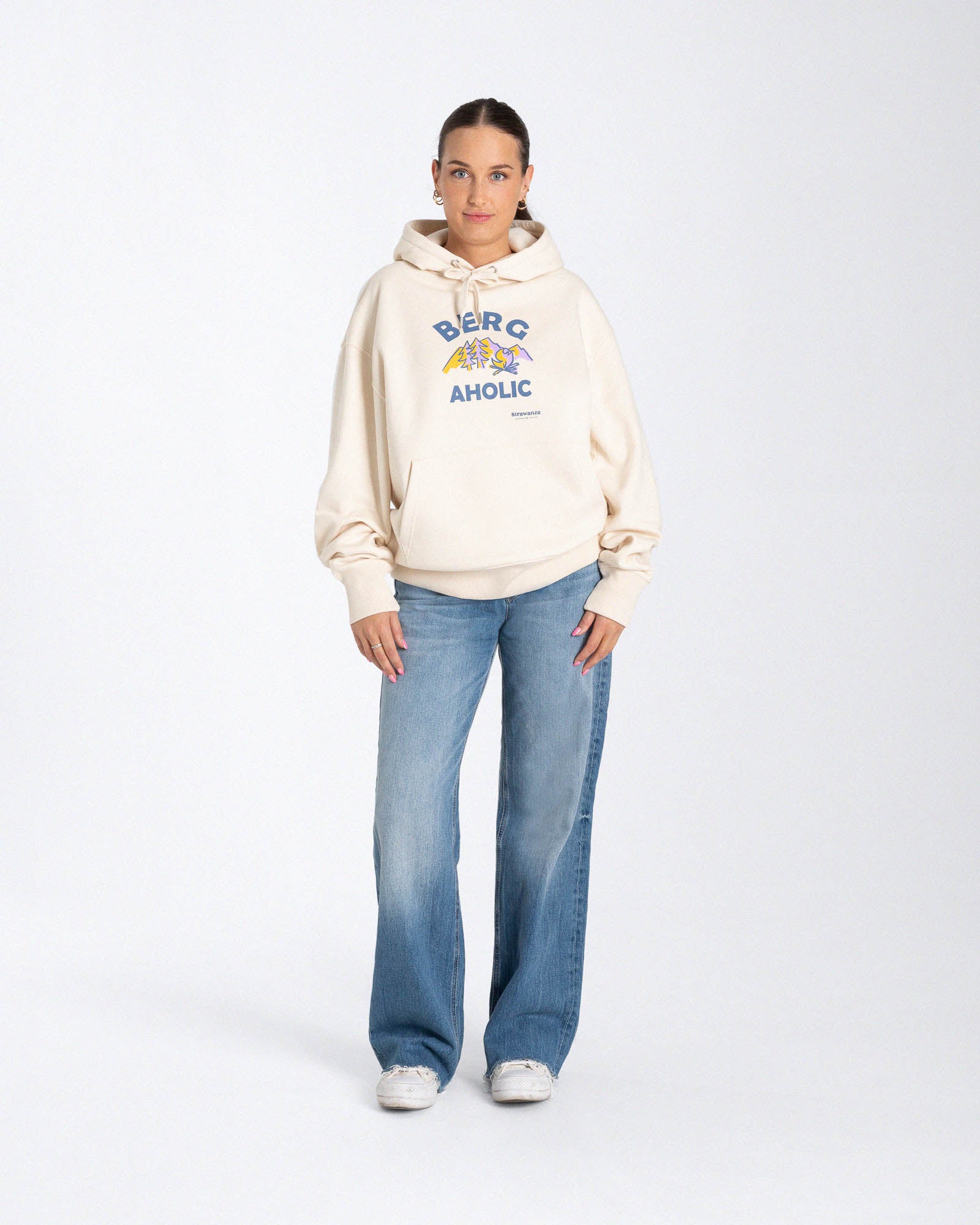 Ein weibliches Model trägt einen Hoodie in der Farbe Natur mit einem Motiv vorne auf der Höhe der Brust, welches aus lila-orangenen Bergen, Bäumen und einem Lagerfeuer  und einem dunkelblauen "BERGAHOLIC" Schriftzug besteht. 