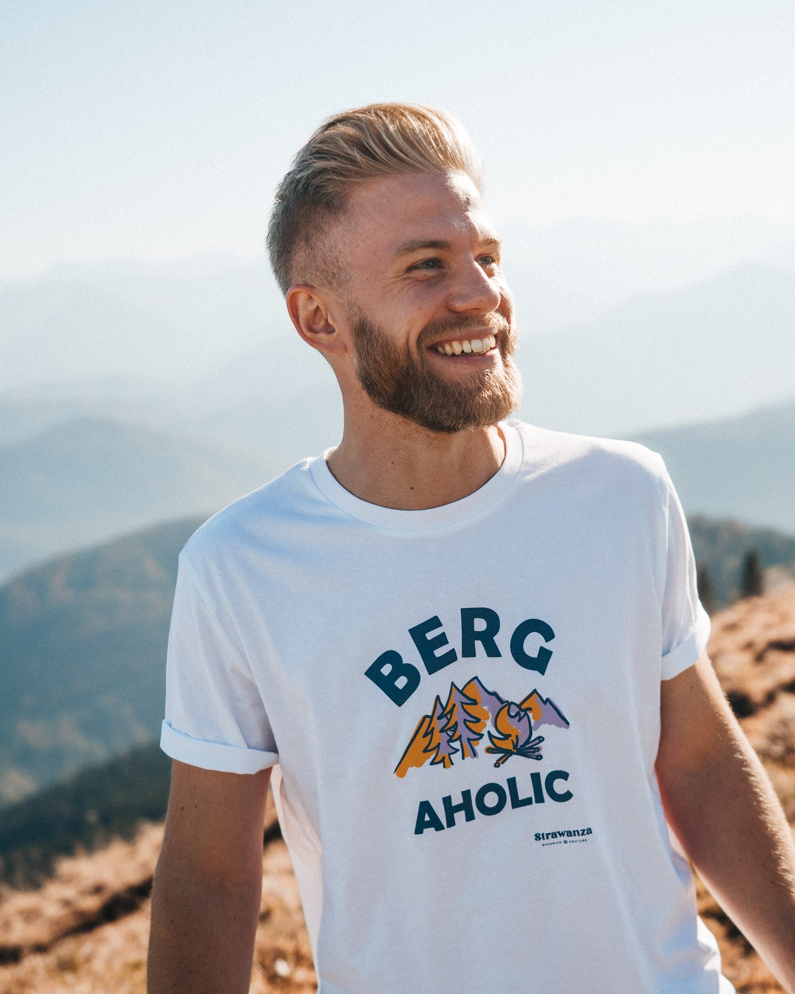 Ein männliches trägt ein weißes T-Shirt mit einem orangen, lilanen Berge und Feuerstelle Motiv, wo zudem noch ein blauer Schriftzug mit dem Wort "BERGAHOLIC" drum herum steht. 