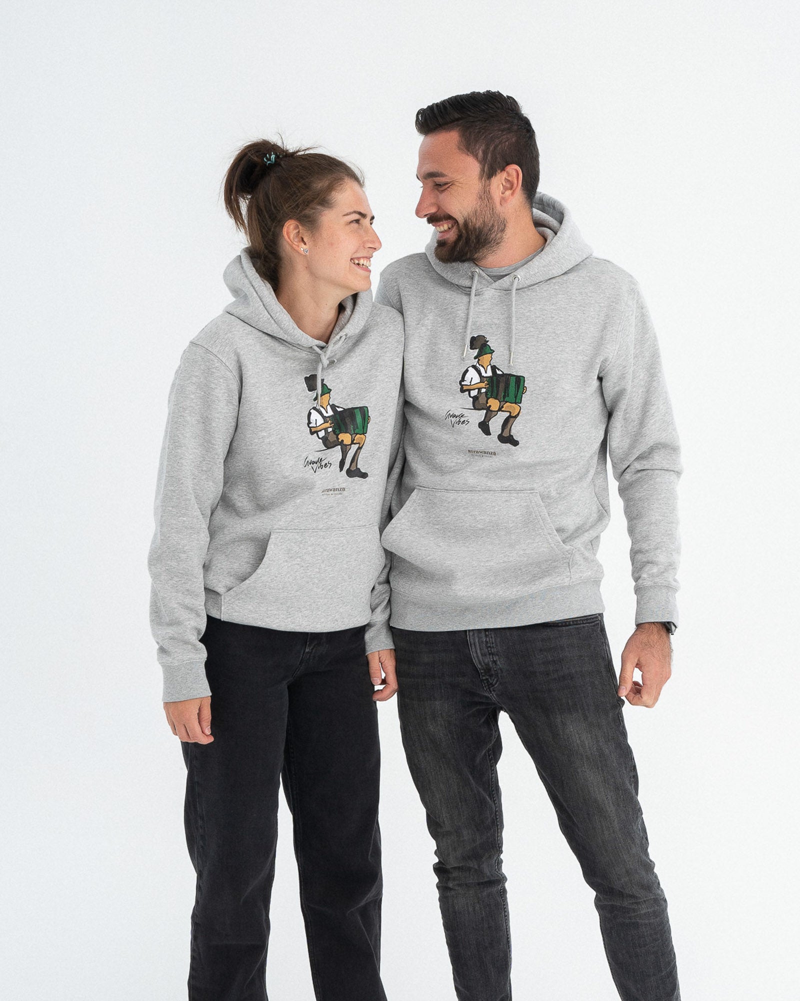 Ein männliches und weibliches Model tragen beide einen grauen Hoodie mit einem Akkordeon spielenden Mann in Tracht und einem  schwarzen "Guade Vibes" Schriftzug.