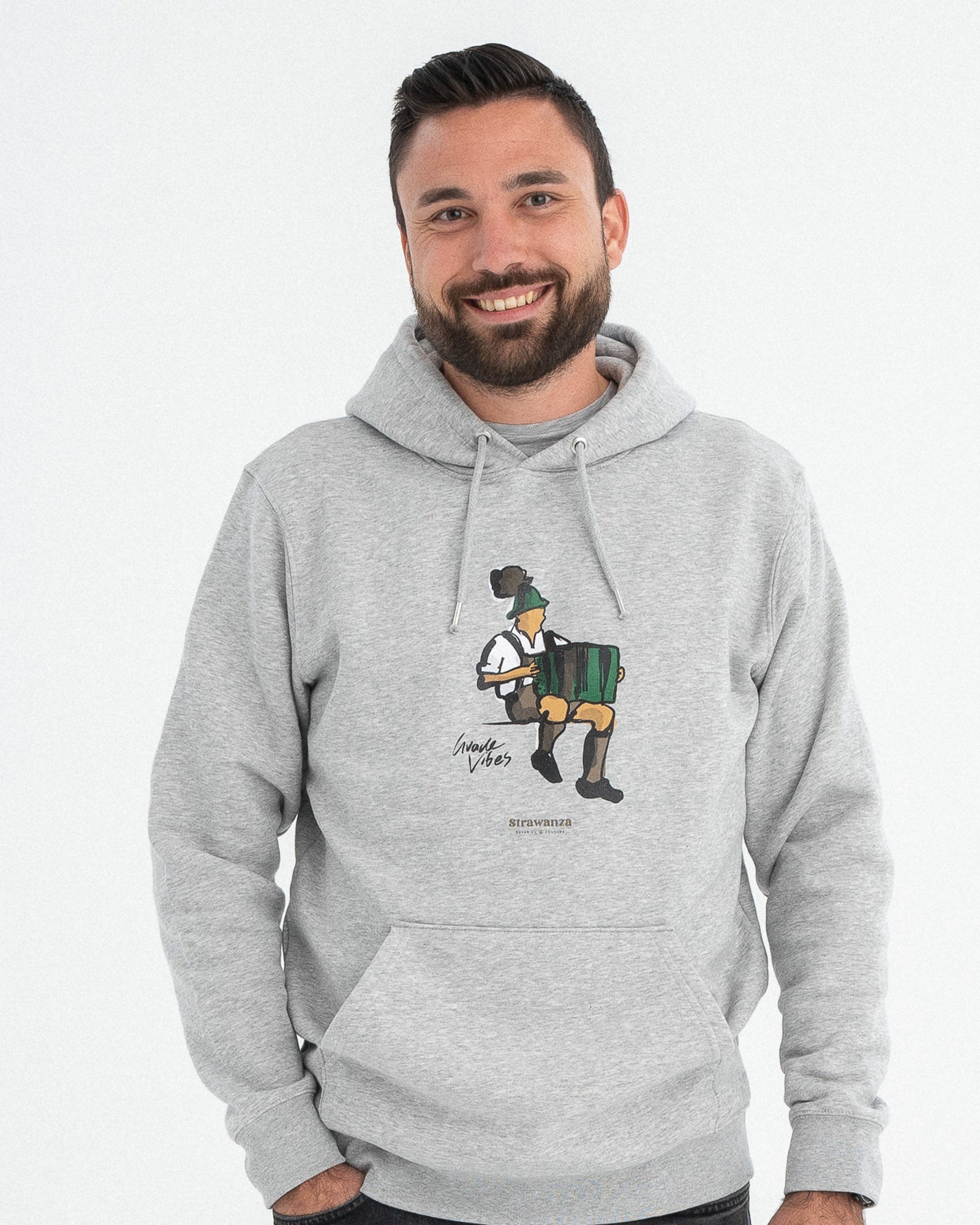 Ein männliches Model trägt einen grauen Hoodie mit einem Motiv welches aus einem Mann besteht der Akkordeon spielt und Tracht an hat und einem in schwarz gedruckten "Guade Vibes" Schriftzug.