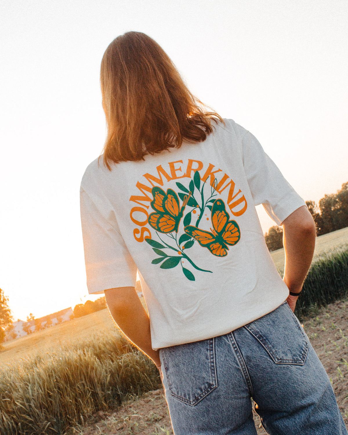 Weibliches Model trägt ein weißes T-Shirt mit einem Schmetterlingsmotiv und einem orangem "SOMMERKIND" Schriftzug als Backprint, in einem Weizenfeld. 