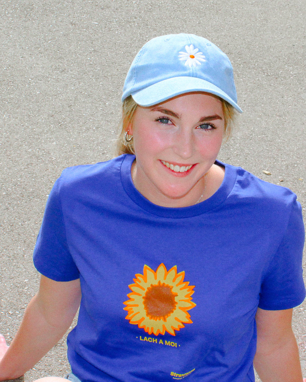 Ein weibliches Model trägt ein blaues T-Shirt mit einer Sonnenblume und einem gelben "LACH A MOI" Schriftzug als Motiv. 