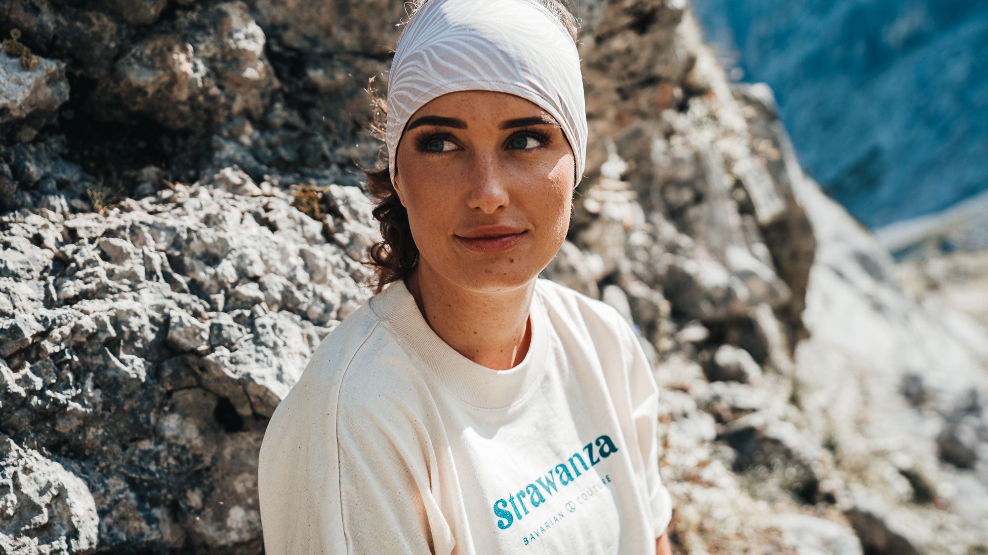 Junge Frau mit Stirnband und weißem T-Shirt der Modemarke Strawanza vor Berglandschaft. 