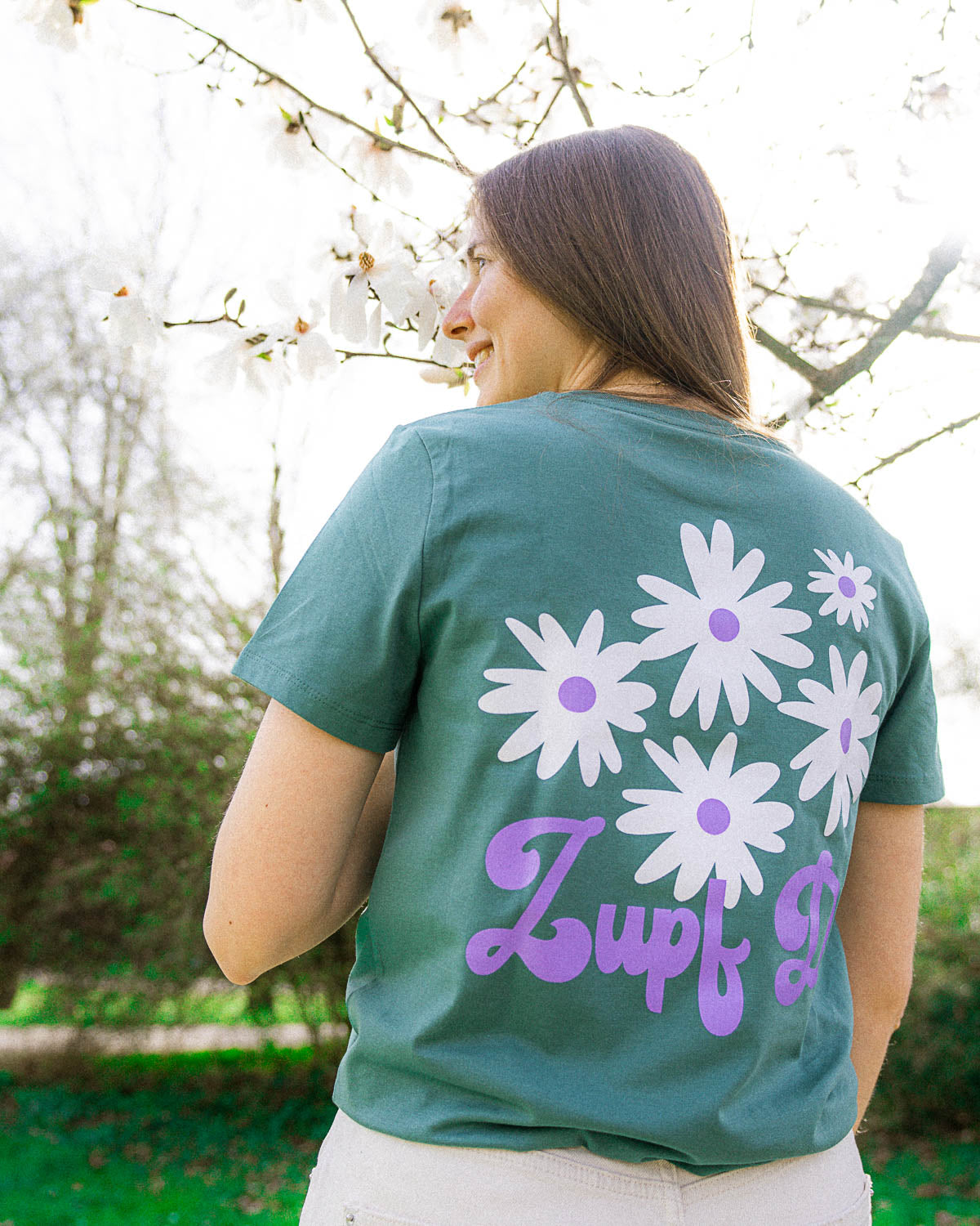 Ein weibliches Model, die in einem Park steht, trägt ein grünes T-Shirt mit weißen Blumen und einem lilanen "Zupf Di!" Schriftzug als Backprint Motiv.