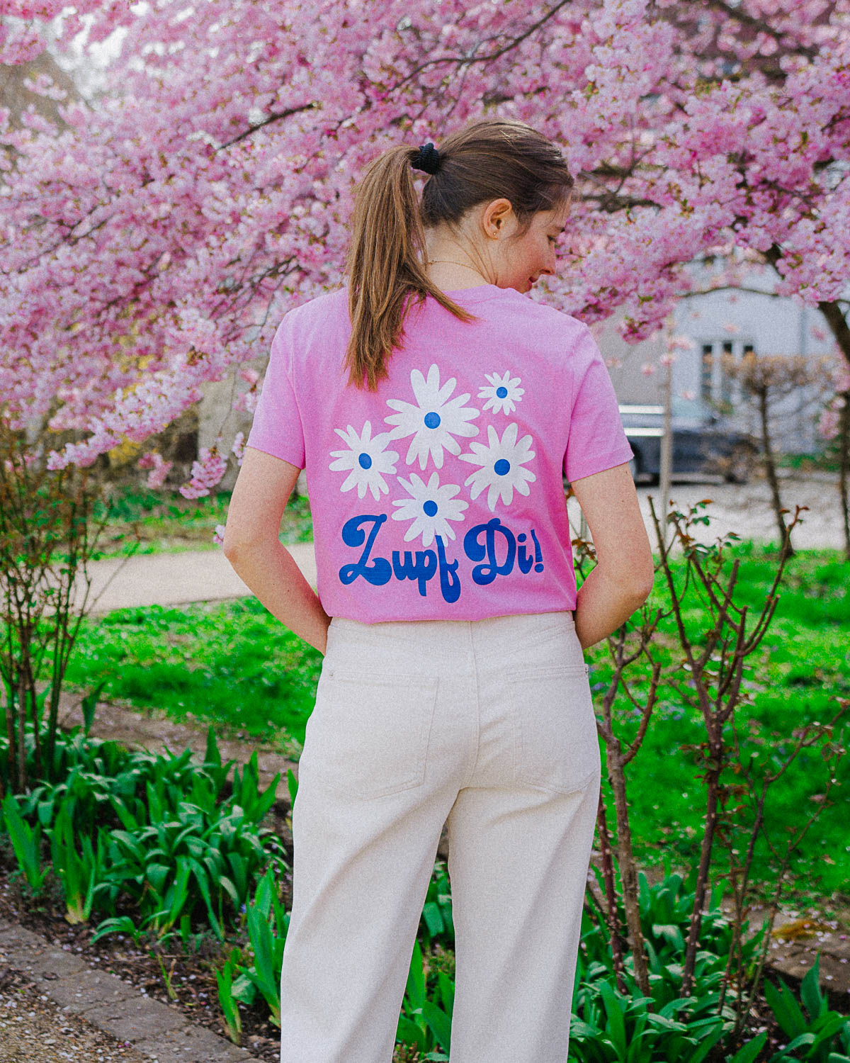 Weibliches Model, die mit dem Rücken zu uns steht,  trägt in einem Park ein pinkes T-Shirt mit einem weißem Blumenmotiv und einer blauen Schrift darunter. 