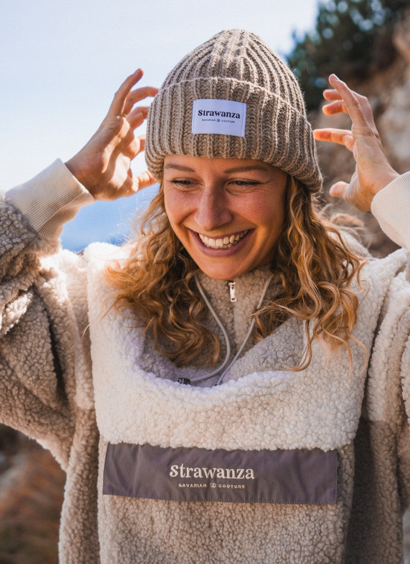 Weibliches Model trägt Sherpa Fleece Anorak von Strawanza vor Alpenkulisse. Sie trägt außerdem eine Wollmütze aus Alpaka Wolle und lächelt. 